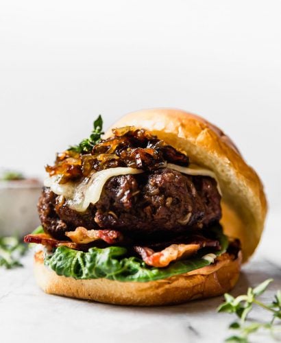 Side angle of French onion soup burger on a brioche burger bun. The burger is topped with lettuce, bacon, white cheddar cheese & caramelized onions. The burger is on a white marble surface, surrounded by a few sprigs of fresh thyme.