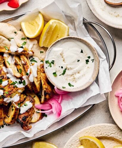 An overhead shot of garlic yogurt shawarma sauce in a small bowl alongside chicken shawarma in a metal tray. The tray is surrounded by plates of lemon wedges, pickled red onions and tomato and a large bowl of sauce on a white textured surface.