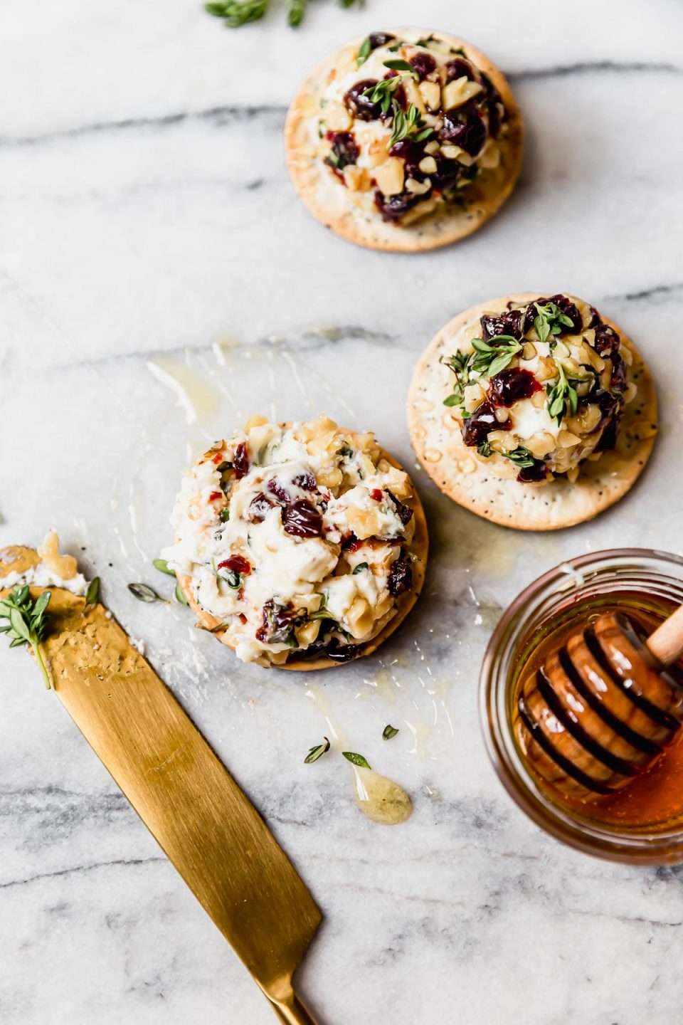 3 goat cheese truffles, coated with chopped nuts & tart cherries, served on crackers & topped with honey. One goat cheese ball is smashed, slightly. A gold knife & small bowl of honey surround the truffles.
