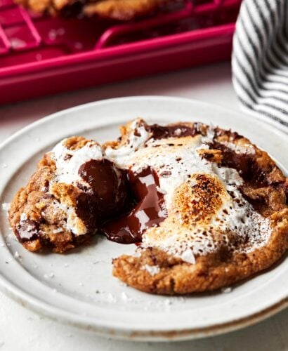 A close-up side shot of a baked s'mores cookie with melted chocolate and marshmallow fluff oozing from the center. The cookie is on a small white plate on a white surface, alongside a striped cloth and a pink baking sheet of cookies.
