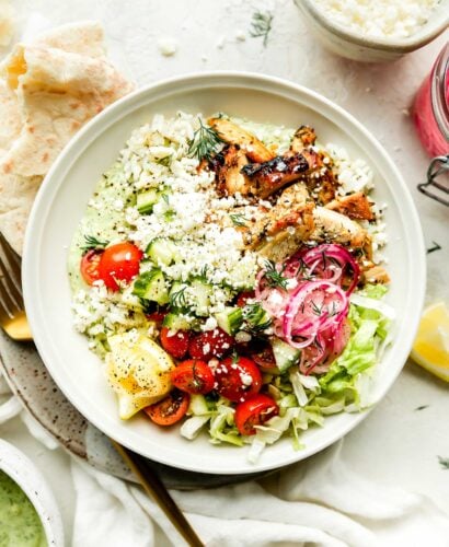 An assembled greek chicken bowl sits on a small plate atop a white surface with a white cloth. A pita and gold fork rest alongside the bowl, and a bowl of avocado tzatziki is on the counter beside it. A bowl of feta, jar of pickled onions, fresh dill, & lemon can be seen in the background.