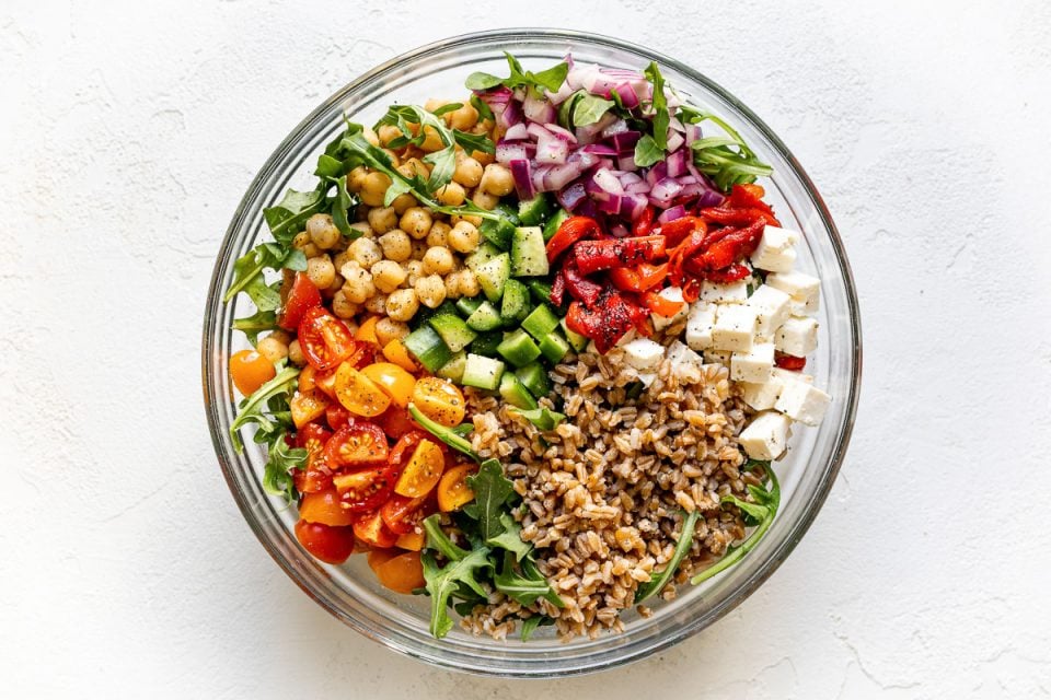 Greek farro salad in large clear glass bowl atop a white surface. The farro salad ingredients are grouped in the bowl - lemony red onions, chickpeas, cucumber, tomatoes, farro, feta, etc.
