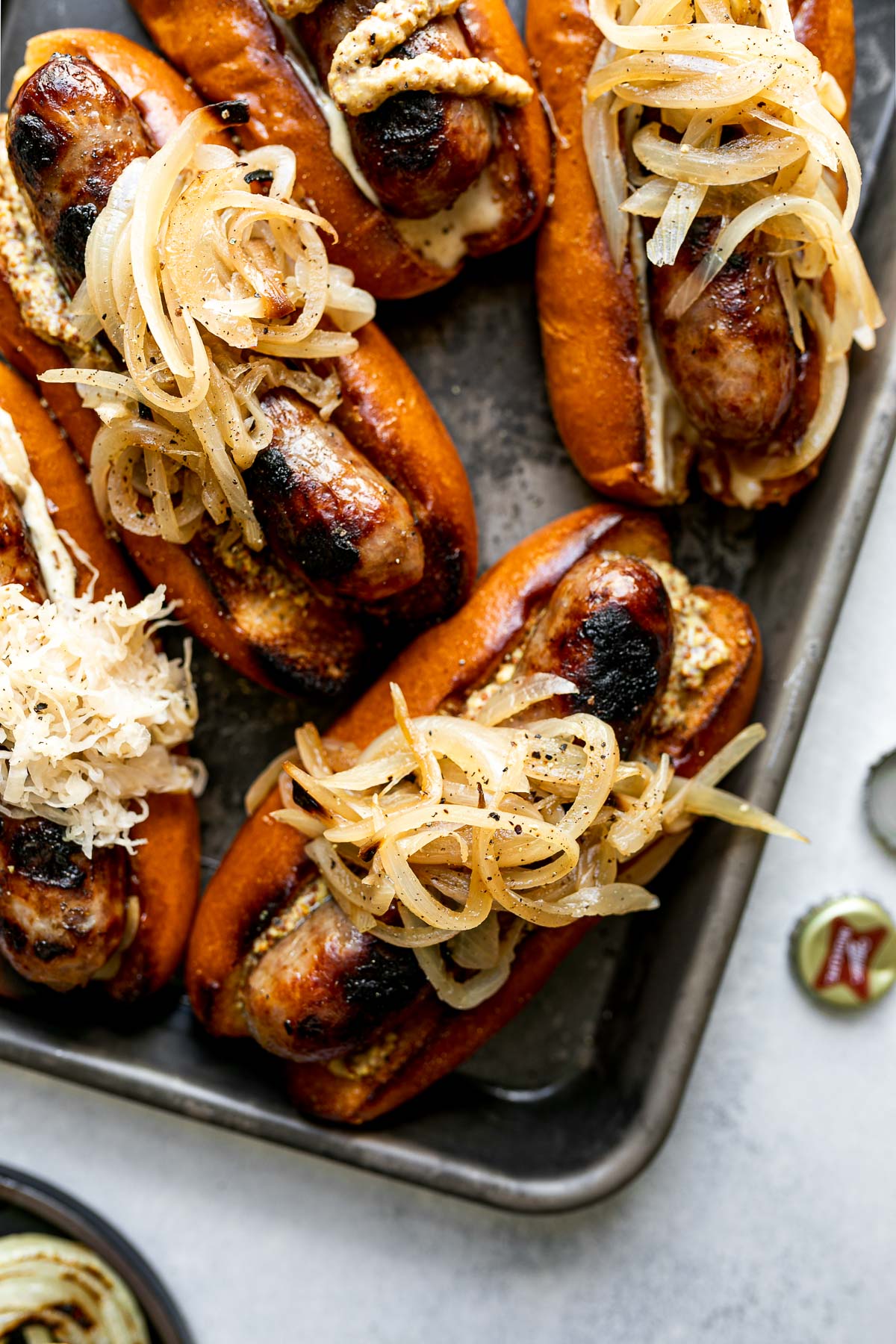 Grilled Wisconsin Beer Brats arranged on a small Nordicware baking sheet. The brats sit on toasted brioche buns, topped with mayonnaise, mustard, braised onions, sauerkraut, etc. The baking sheet sits atop a light blue surface.