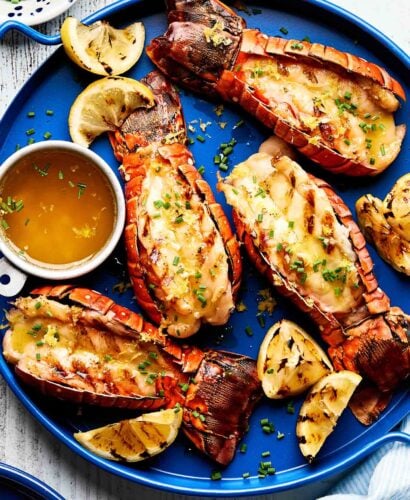 An overhead shot of grilled lobster tails garnished with chives on a bright blue plate. Lemon wedges and a small dish of melted butter sit alongside the tails.