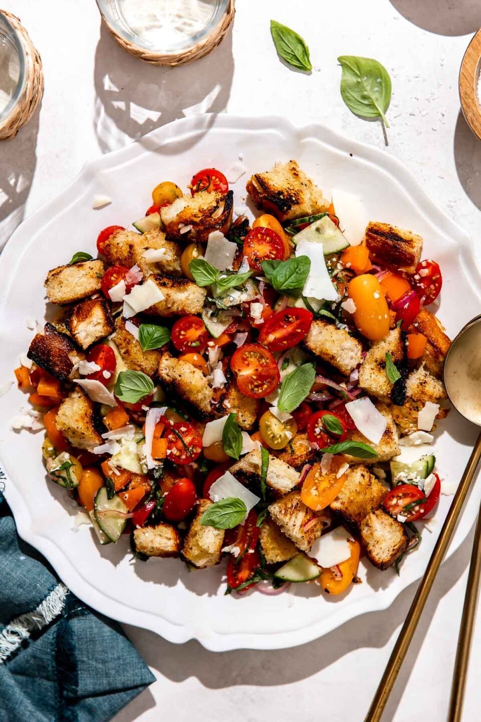 Grilled panzanella salad is served atop a white serving platter that sits atop a white textured surface. The panzanella salad has been garnished with fresh herbs and shaved asiago cheese. Two gold serving spoons rest atop the platter and the platter is surrounded by two drinking glasses, a small pinch bowl filled with kosher salt, and a blue linen napkin.
