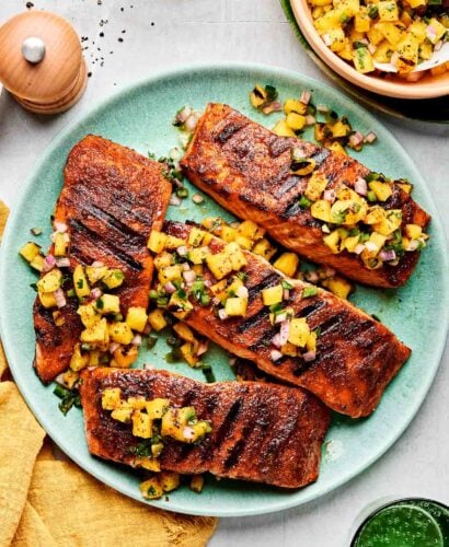 Four grilled salmon fillets topped with pineapple salsa rest atop a light blue plate that sits atop a light gray surface. A bowl of pineapple salsa, a pepper mill, a yellow linen napkin, and a green drinking glass surround the plate at center.