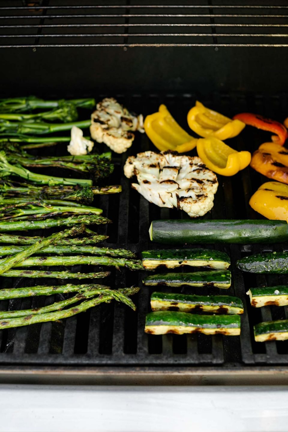A straight on shot of a variety of grilled veggies including grilled broccolini, grilled asparagus, grilled cauliflower, grilled zucchini, grilled onion, & grilled peppers that are being grilled on gas grill grates. Many of the veggies have grill marks on them after being flipped to the other side.
