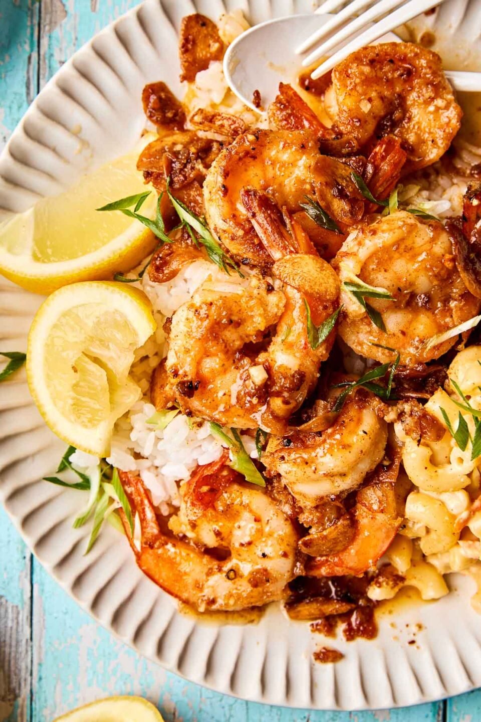 A plate of seasoned shrimp on white rice, garnished with green onions, served with lemon slices and macaroni and cheese on a white, ridged plate.