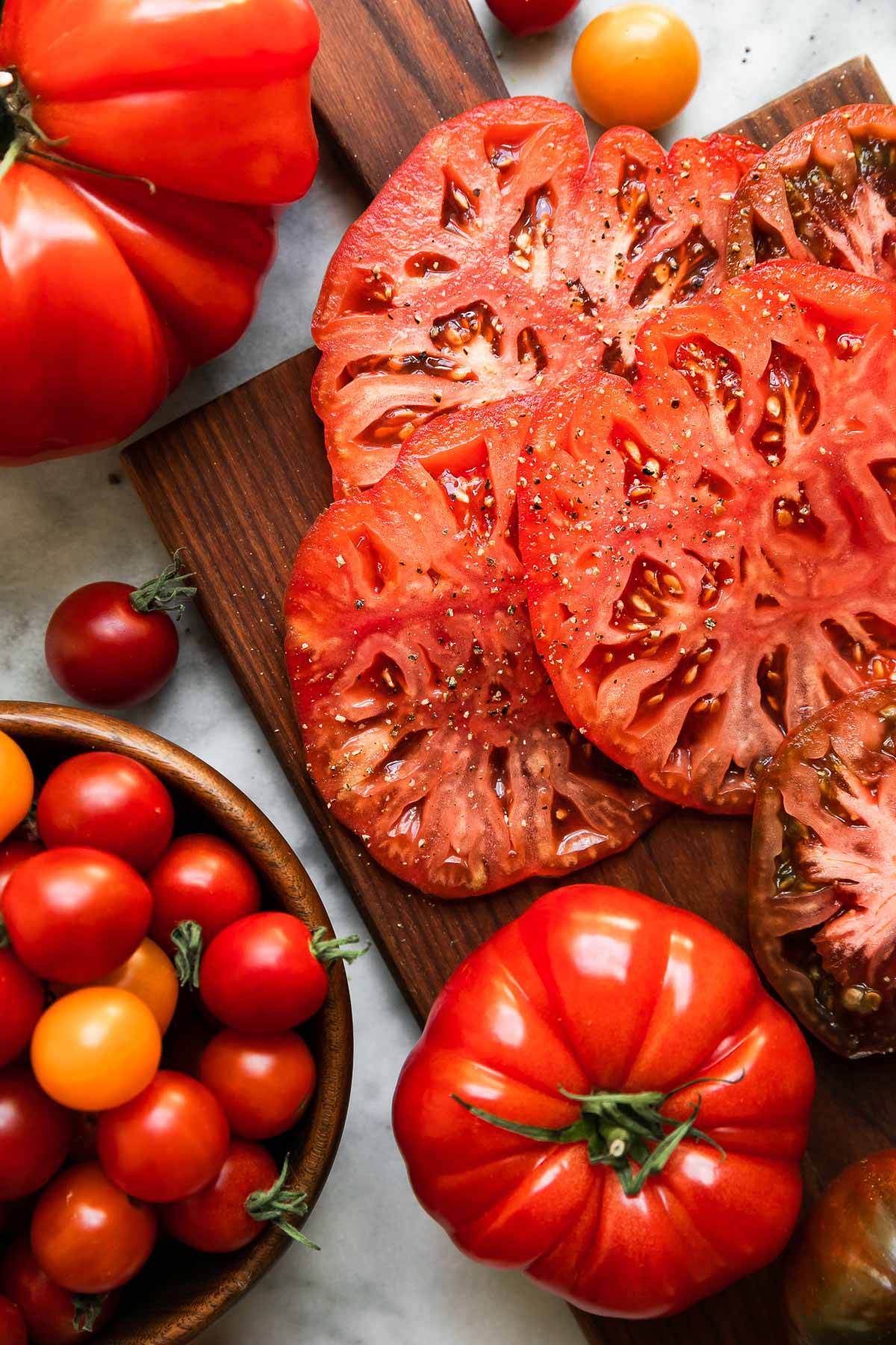 Whole and sliced heirloom tomatoes atop a small wooden cutting board and whole cherry tomatoes arranged in a small wooden bowl sit atop a white and gray marble surface. These tomatoes are prepped for a tomato burrrata salad.