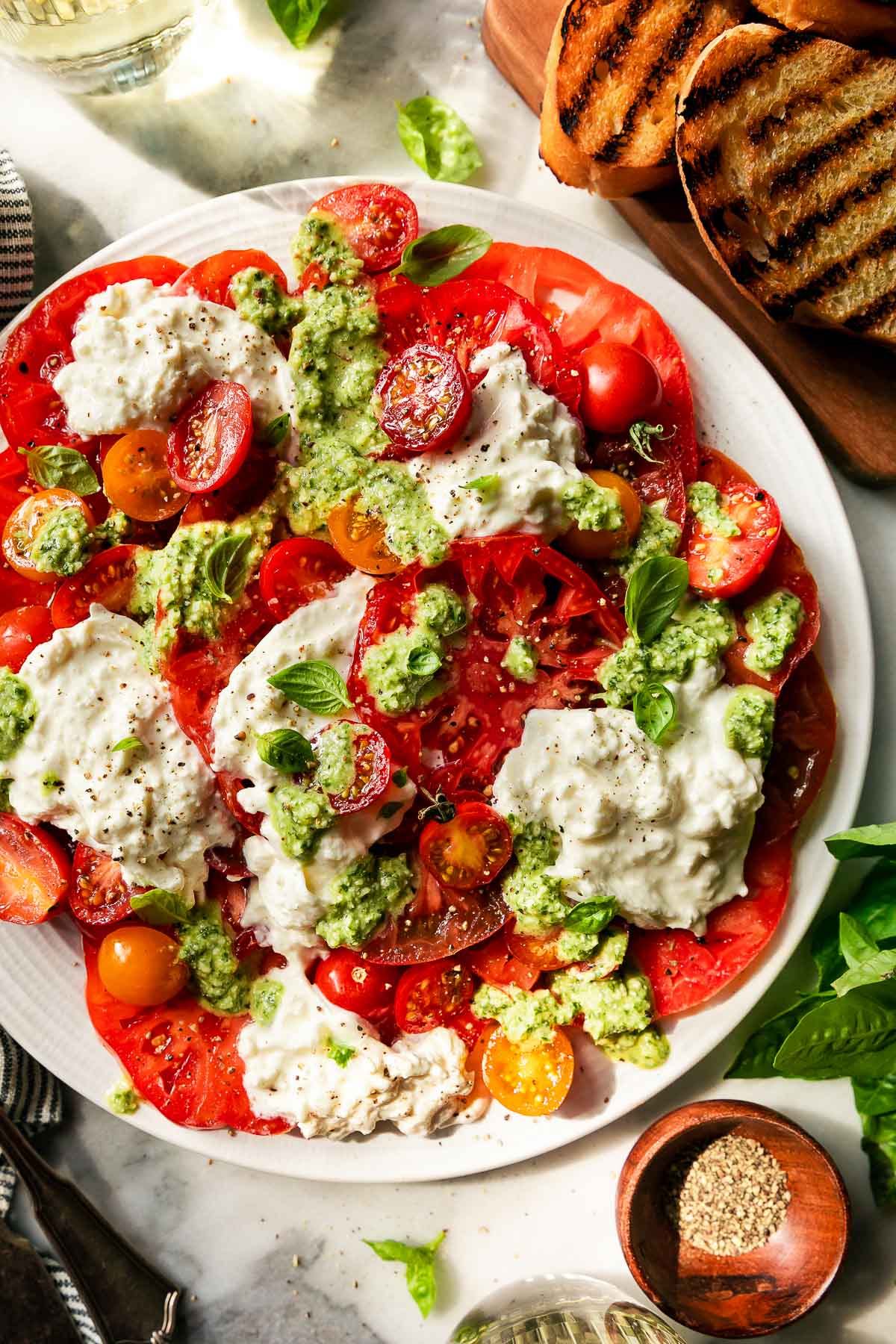 Tomato and burrata salad arranged on large white serving platter sits atop a white and gray marbles surface. The platter is surrounded by a wooden serving board topped with grilled bread, fresh basil, a small wooden pinch bowl filled with ground black pepper, two small drinking glasses filled with white wine, a gray and white striped linen napkin, and silver servingware.