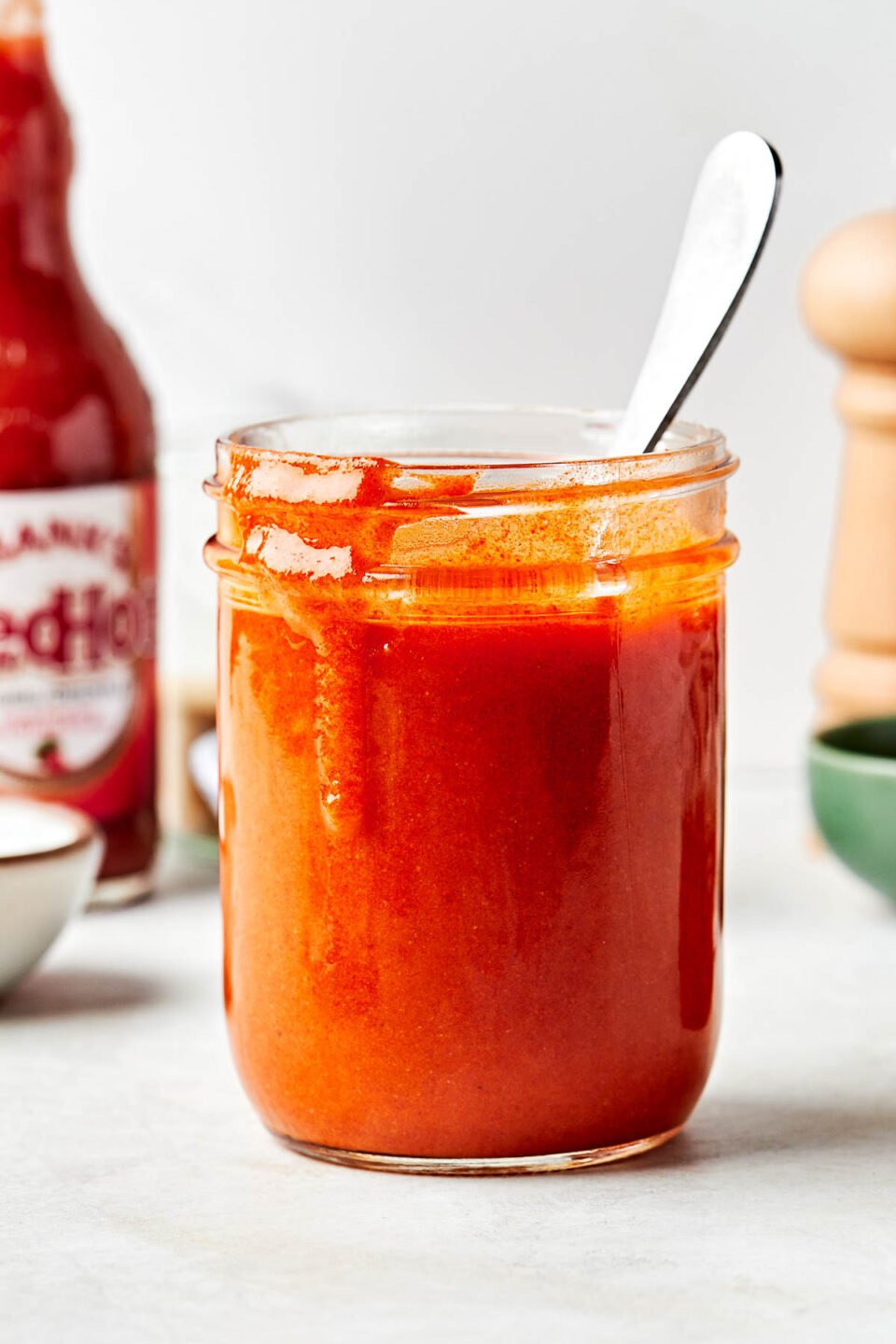 A side shot of a glass jar of homemade buffalo sauce sitting on a white surface alongside a bottle of Frank's Red Hot, small bowls, and a pepper grinder.
