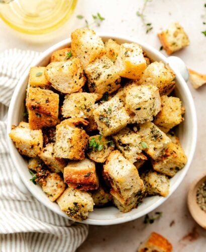 An close-up overhead shot of homemade croutons in a white bowl atop an off-white surface. A striped cloth, bowl of pepper, and a jug of olive oil sit beside the bowl.