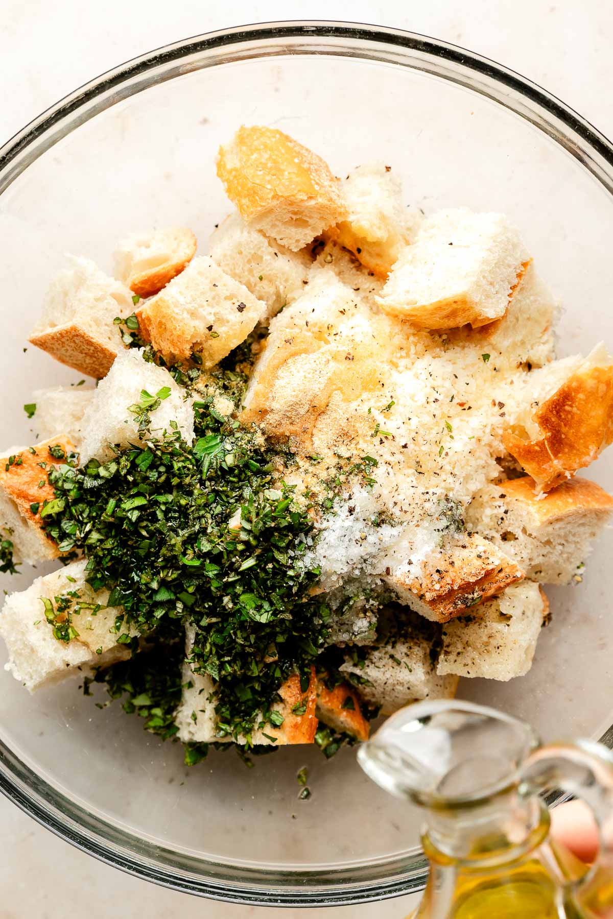 An overhead shot of ingredients in a glass bowl atop an off-white surface: cubed bread, chopped herbs, salt, pepper, and parmesan. Olive oil is being drizzled from a glass jug over the ingredients.
