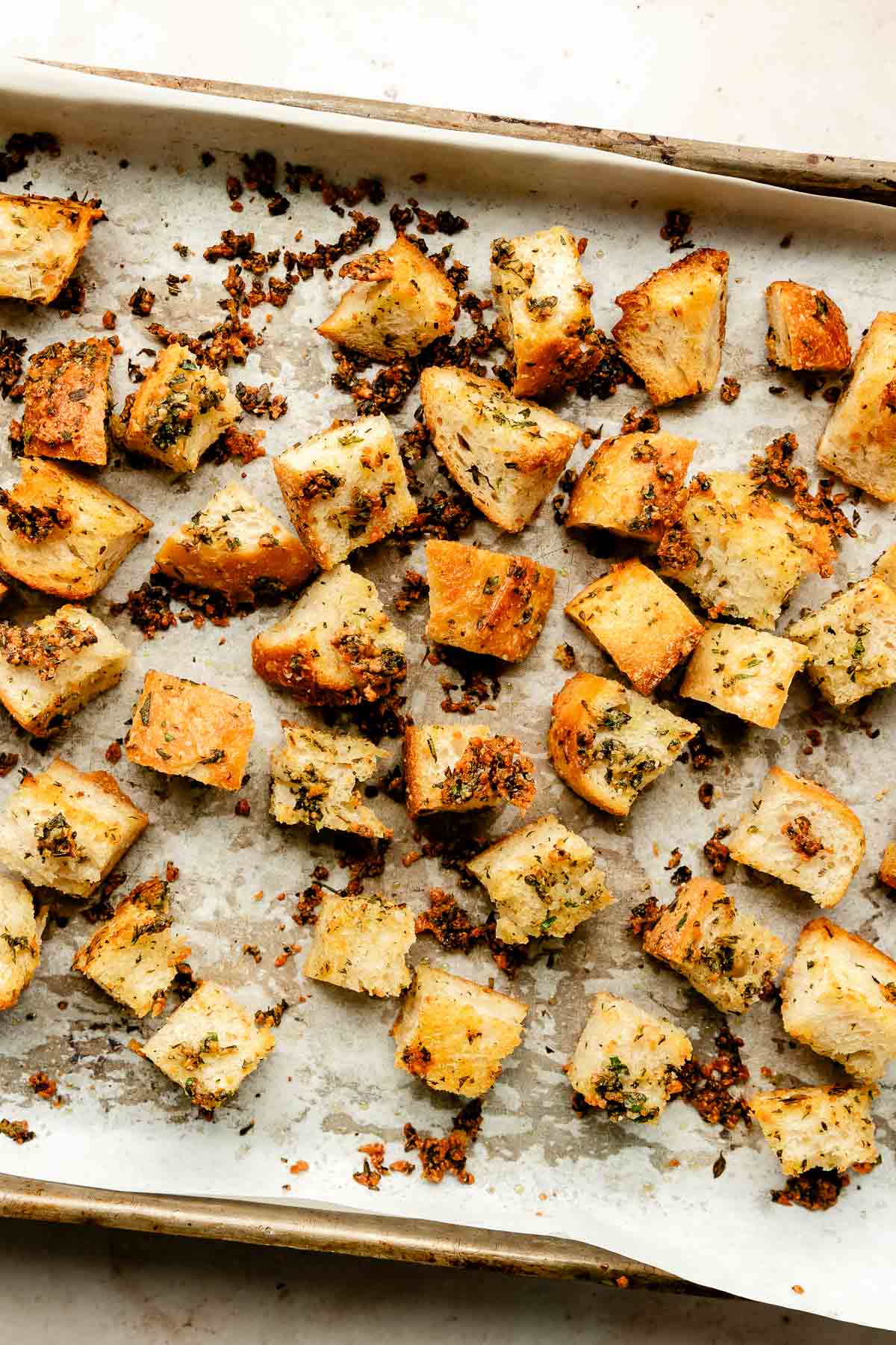 An overhead shot of baked garlic herb croutons on parchment paper on a sheet pan atop an off-white surface.