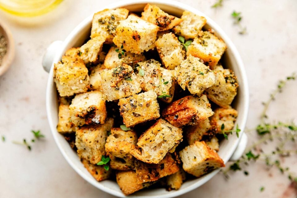 An close-up overhead shot of homemade croutons in a white bowl atop an off-white surface.
