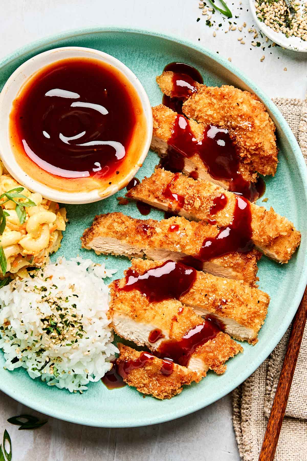 An overhead shot of sauce drizzled over chicken katsu on a light teal plate with white rice and mac salad.