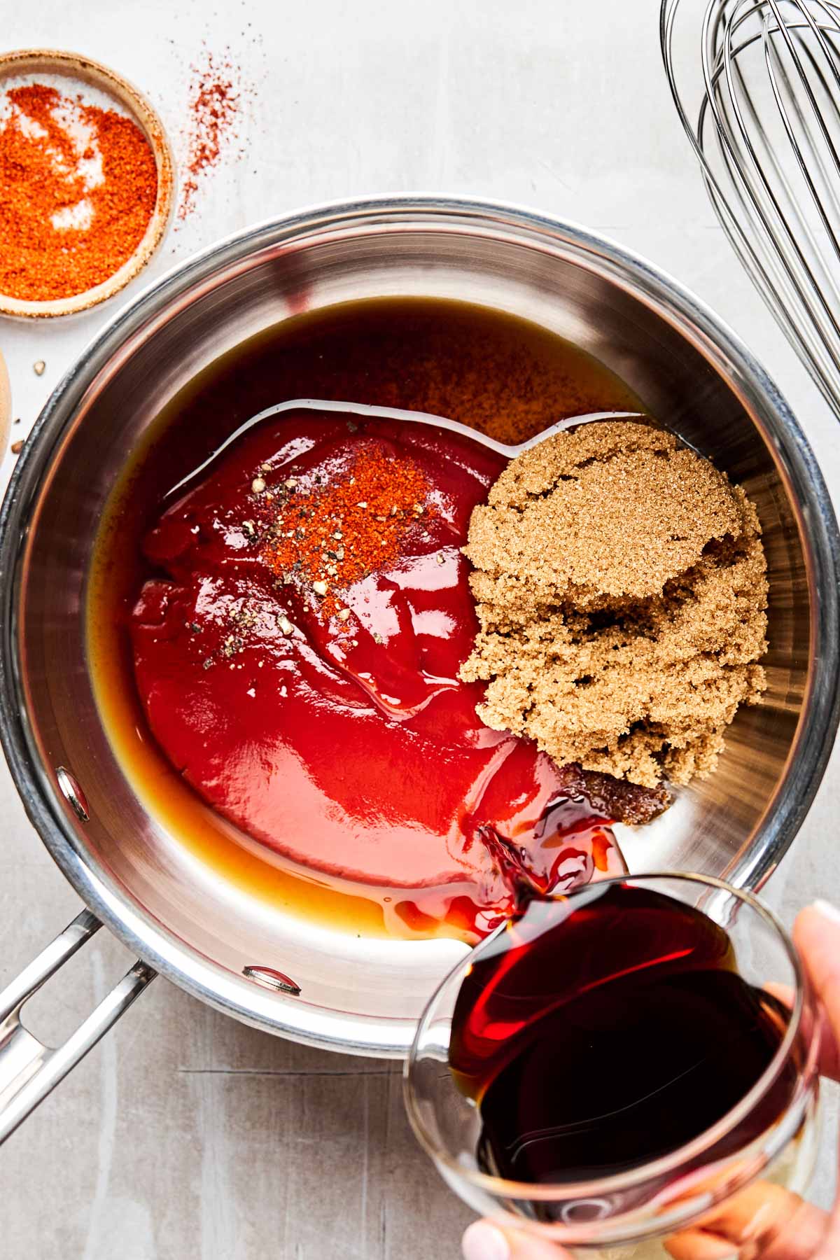 An overhead shot of shoyu being poured into a saucepan with ketchup, Worcestershire sauce, brown sugar, salt, pepper, and cayenne pepper. The saucepan sits on a light grey textured surface.