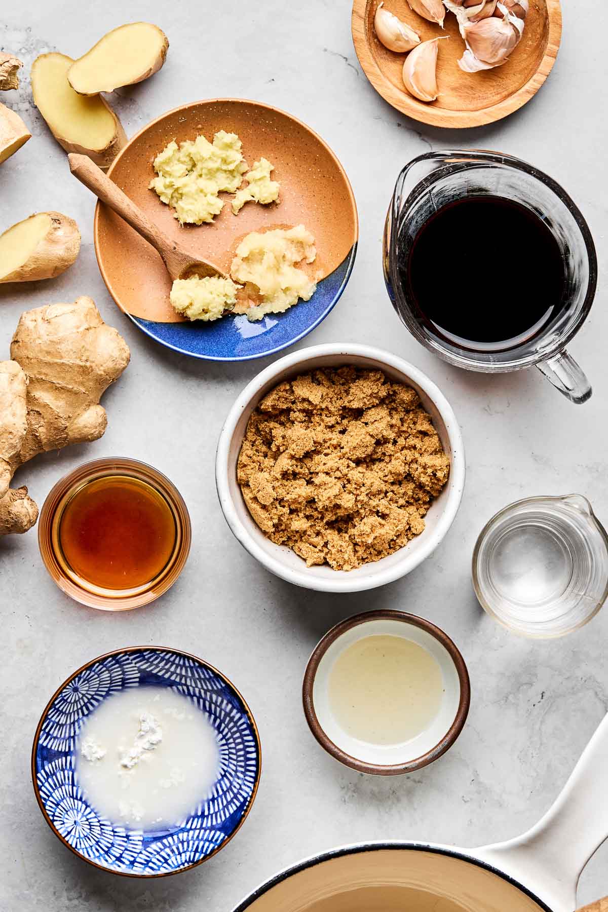 An overhead shot of ingredients displayed in various bowls on a white marbled surface: brown sugar, shoyu, ginger, garlic, water, mirin, toasted sesame oil, and cornstarch slurry.