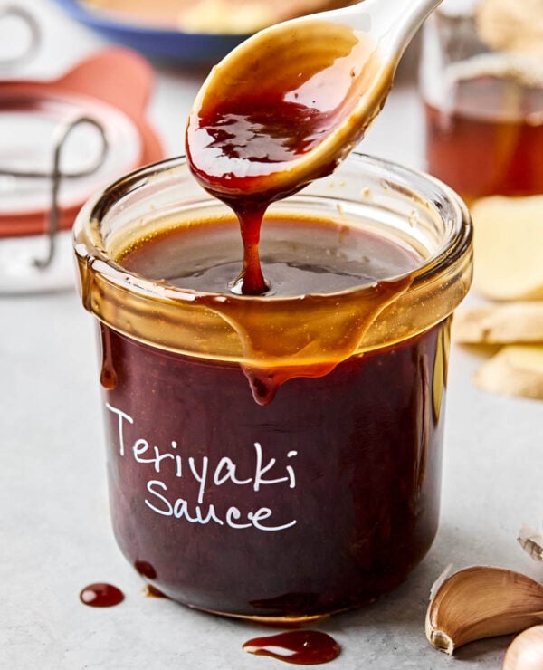 A side shot of a spoon drizzling teriyaki sauce into a labeled glass jar atop a white marbled surface. garlic, ginger, and bowls of ingredients can be seen in the background.