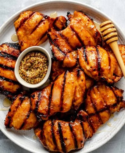 Grilled honey mustard chicken thighs shown on a white speckled plate atop a light blue surface. The chicken is garnished with a drizzled of fresh honey & a small bowl of dijon mustard rests on the plate nestled between the chicken. A wooden honey dipper rests on the side of the plate.