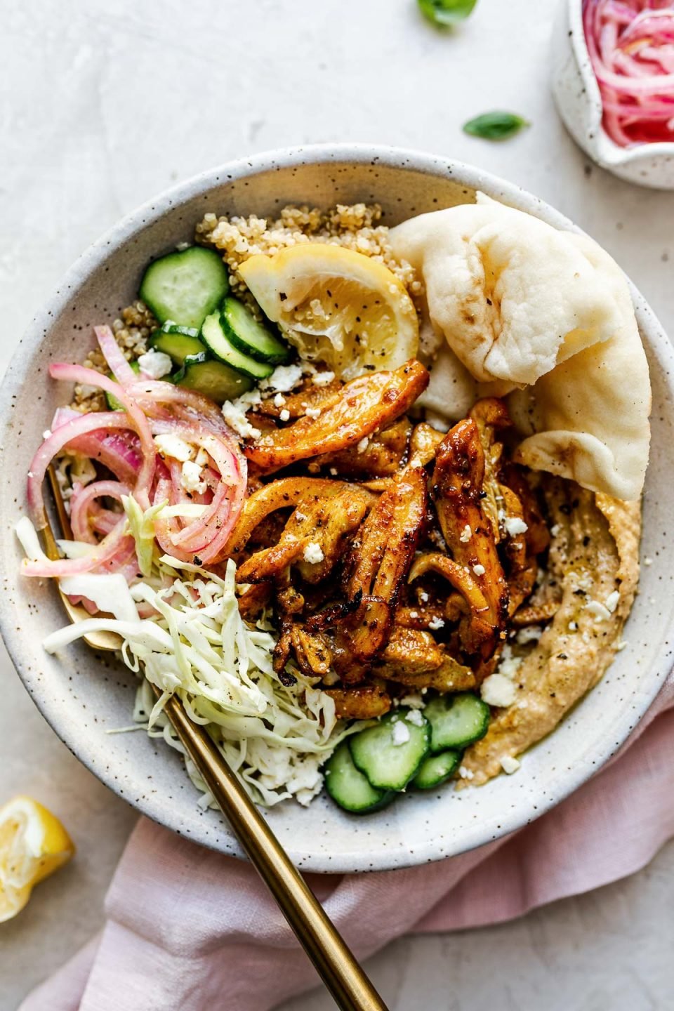 Chicken Shawarma Hummus Bowl with quinoa, cucumbers, pickled red onion, shredded cabbage, & naan bread shown in a speckled gray ceramic bowl. The bowl sits atop a creamy cement surface, surrounded by lemon wedges, fresh basil leaves, a small bowl of pickled red onions, & a pink linen napkin.