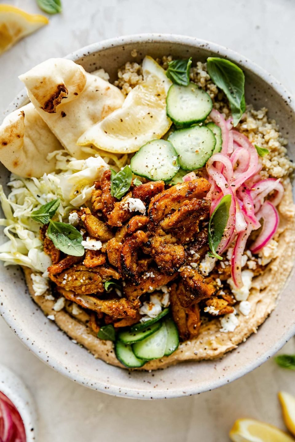 Chicken Shawarma Hummus Bowl with quinoa, cucumbers, pickled red onion, shredded cabbage, & naan bread shown in a speckled gray ceramic bowl. The bowl sits atop a creamy cement surface, surrounded by lemon wedges, fresh basil leaves & a small bowl of pickled red onions.