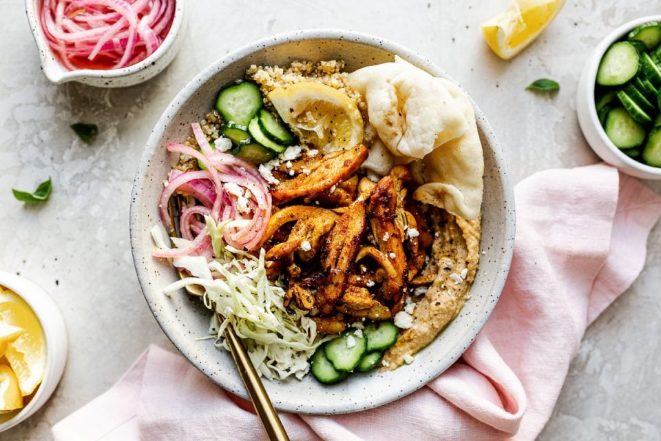 Chicken Shawarma Hummus Bowl with quinoa, cucumbers, pickled red onion, shredded cabbage, & naan bread shown in a speckled gray ceramic bowl. The bowl sits atop a creamy cement surface, surrounded by lemon wedges, fresh basil leaves, a small bowl of pickled red onions, a small bowl of sliced cucumbers, & a pink linen napkin.