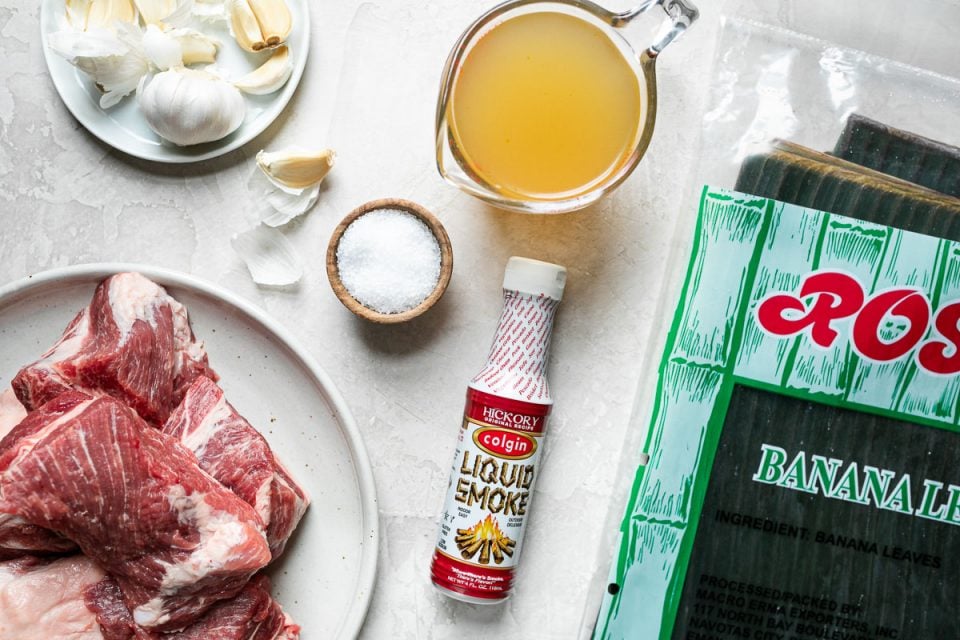 Pork shoulder, garlic, Hawaiian sea salt, Colgin liquid smoke, chicken stock, & a package of frozen banana leaves sit atop a creamy cement backdrop.
