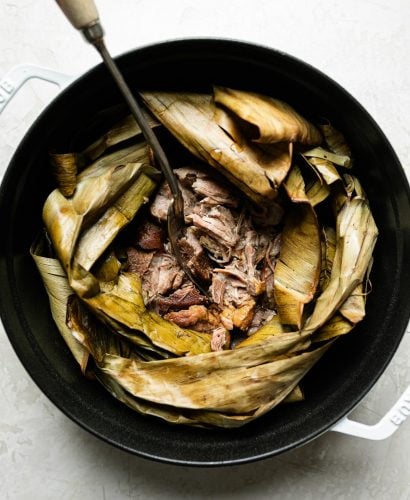 Shredded Kalua pig shown inside dried banana leaf in a white Dutch oven atop a creamy cement surface. A serving fork with a long wooden handle is inserted in the center of the pork.