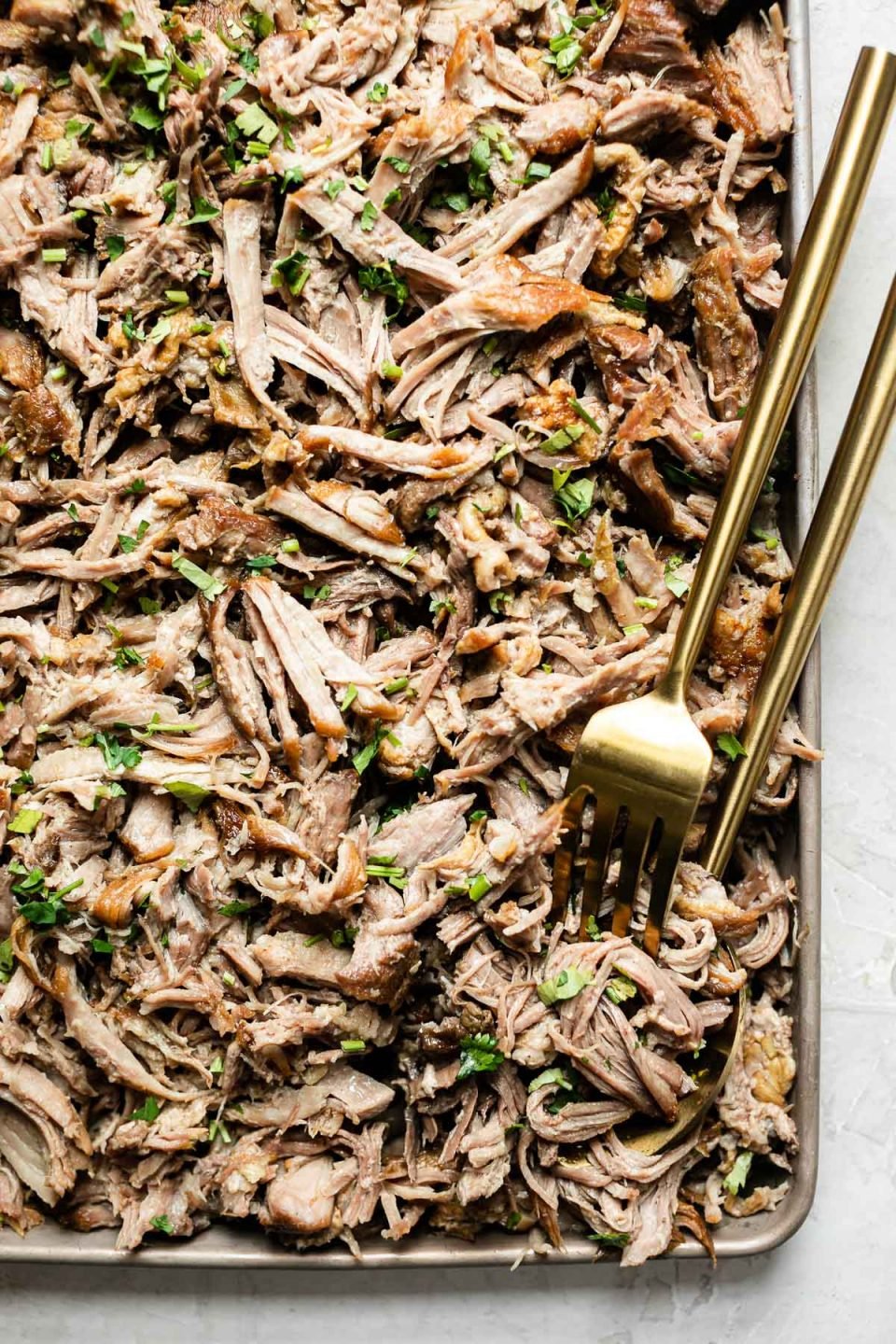 Close up of shredded kalua pork on a small baking sheet, topped with herbs. A gold serving fork & spoon are nestled in the pork.