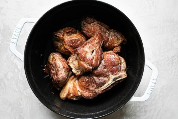 Browned pork shoulder in a white Dutch oven atop a creamy cement backdrop.