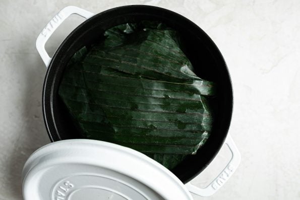 Banana leaf-wrapped pork shown in a white Dutch oven atop a creamy cement surface.