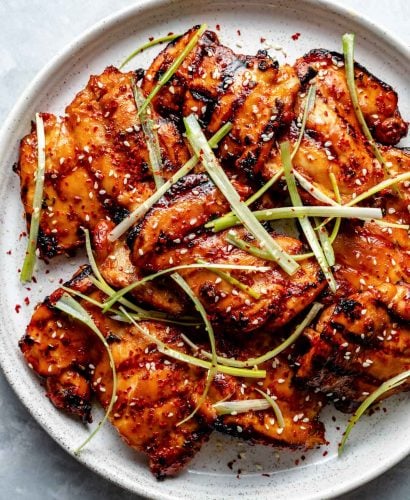 Grilled Korean bulgogi chicken thighs shown on a white speckled plate atop a light blue surface. The chicken is garnished with sesame seeds, shaved green onion, & chili flakes.