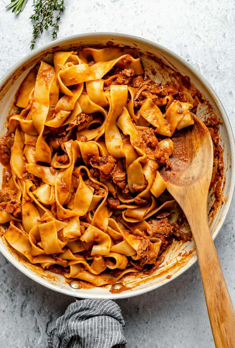Lamb ragu pappardelle in a small skillet atop a light blue surface. A wooden spoon is nestled into the pasta. Next to the skillet are some fresh herbs.