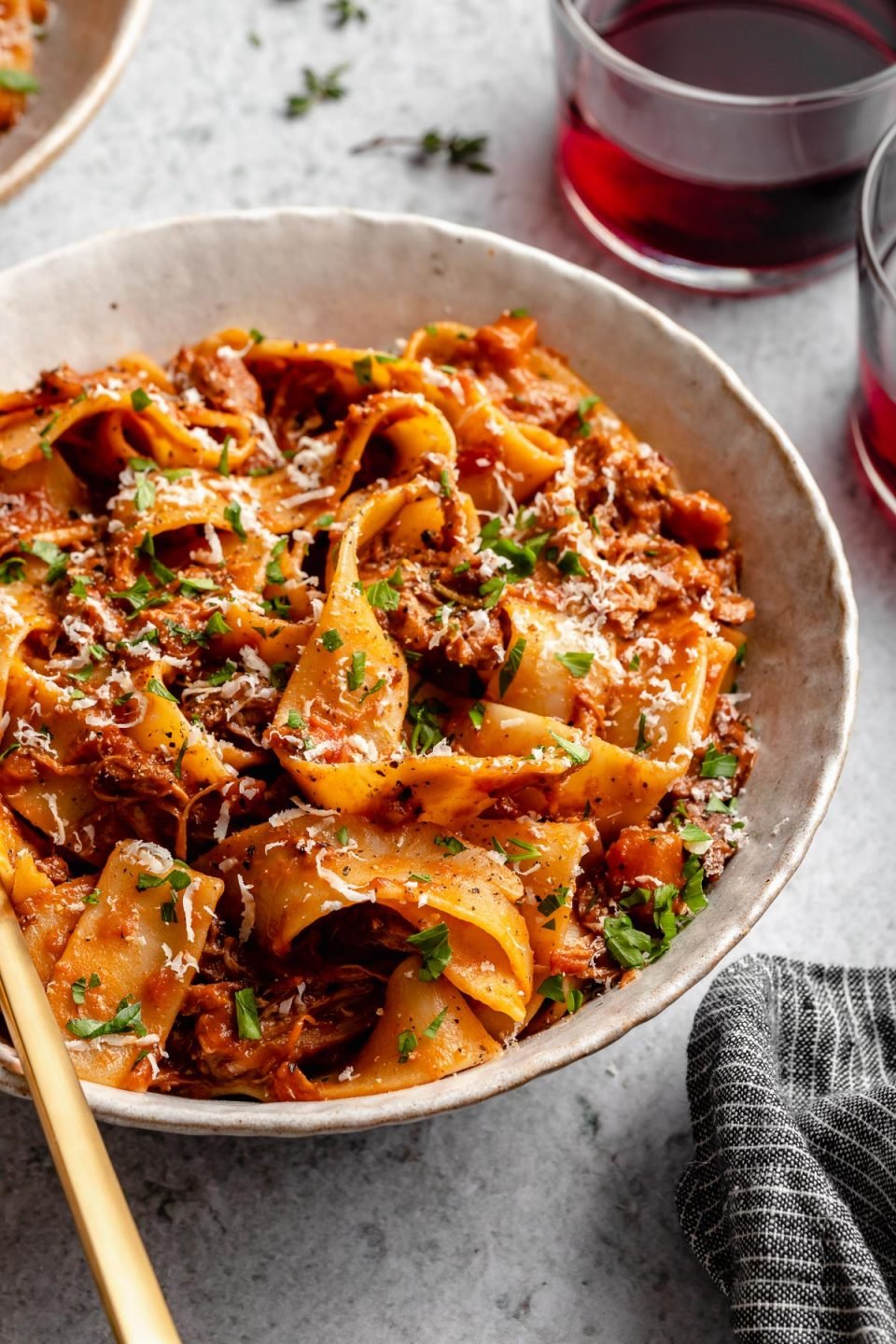 Side angle of Braised Lamb Ragu Pappardelle shown in pasta bowls, topped with grated parmesan and fresh parsley. The bowls sit atop a light blue surface next to fresh herbs, 2 glasses of red wine & a grey striped linen napkin.