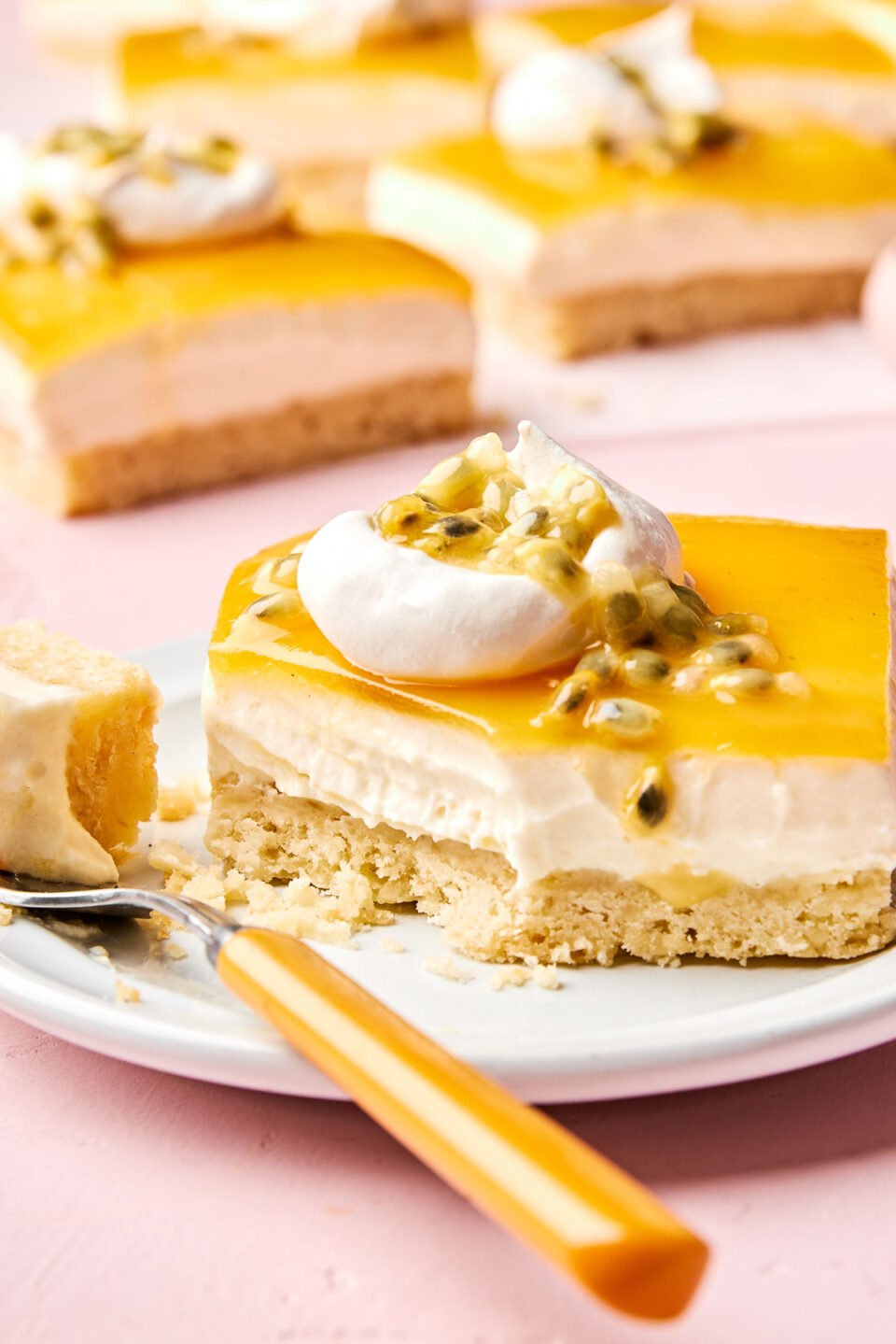 A close-up angled shot of a passionfruit cheesecake bar on a small white plate on a light pink surface. The bar has a bite removed and resting on a fork on the plate. Several more bars sit in the background.