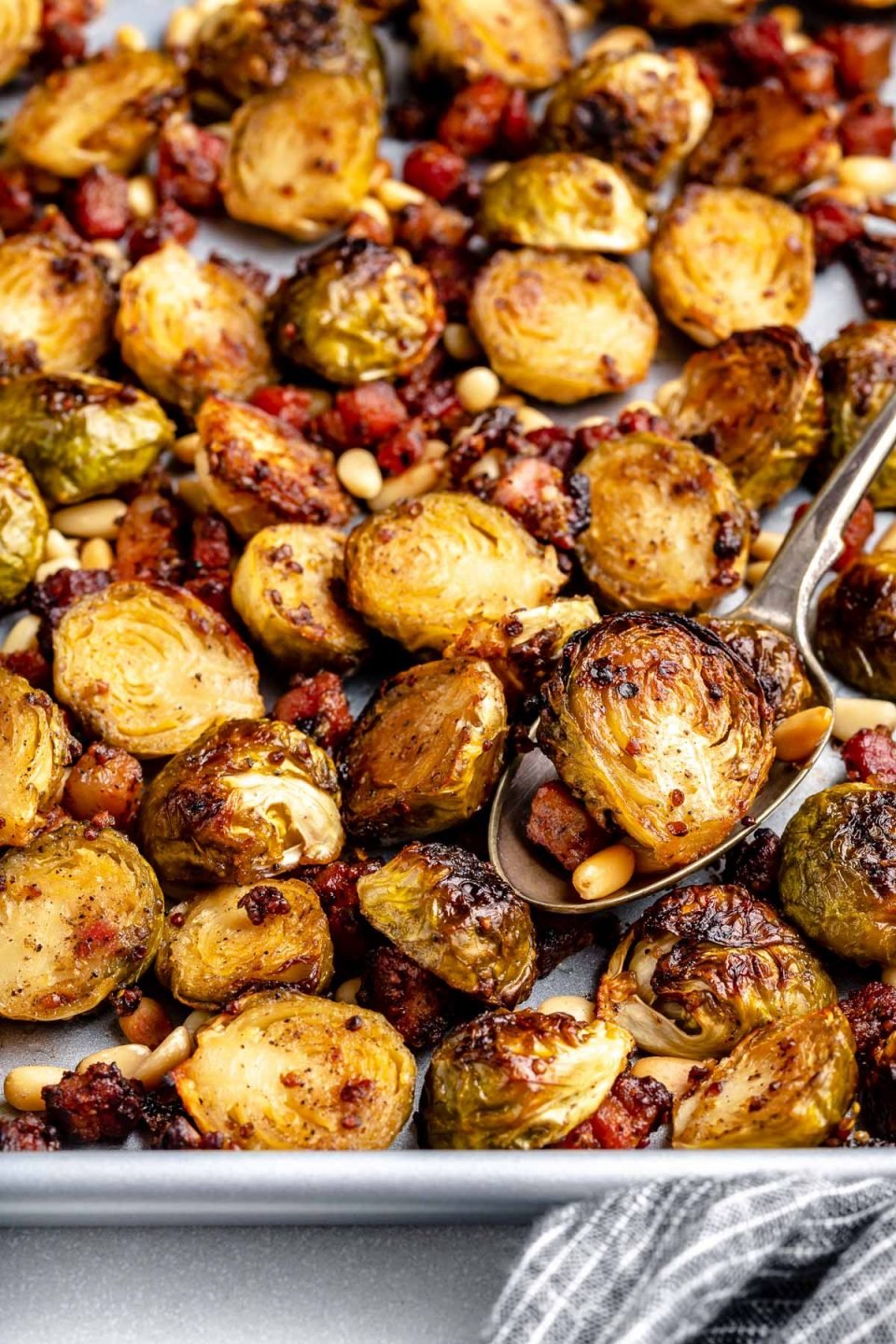 Close view of maple mustard roasted brussels sprouts with pancetta and pine nuts on a large baking sheet. The baking sheet sits atop a light blue surface next to a striped blue linen napkin.