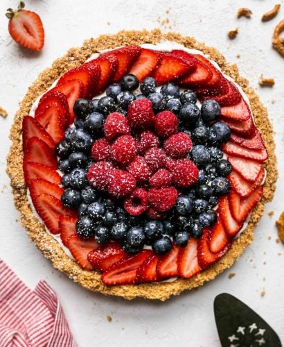 Close up of no-bake summer berry cheesecake tart with pretzel graham cracker crust. The tart is on a white textured surface, topped with fresh strawberries, blueberries, raspberries, & a sprinkle of powdered sugar. Surrounding the tart are loose berries, whole and crumbled pretzels, graham cracker crumbs, a white and red linen napkin, & a pie server utensil.