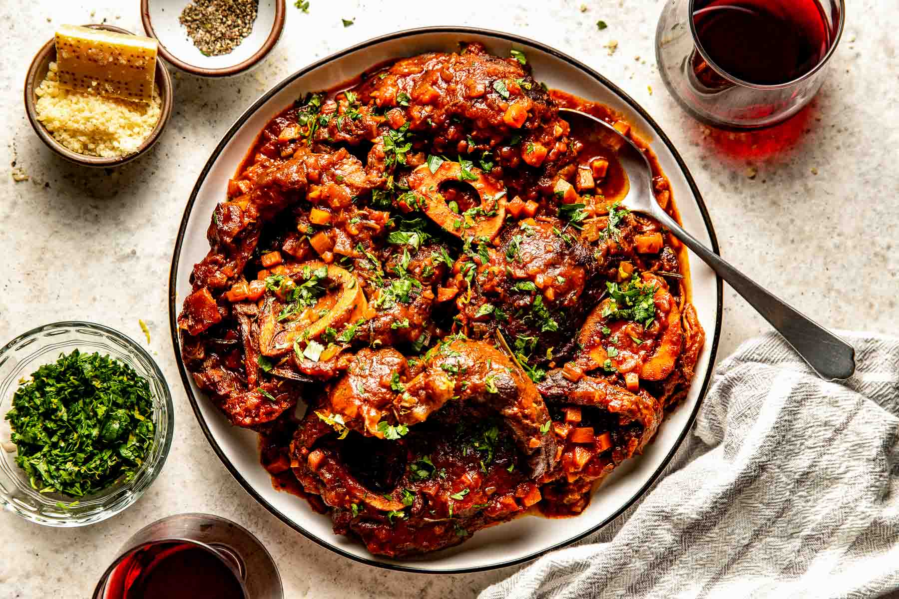 An overhead shot of prepared osso buco garnished with parsley on a white plate atop an off-white textured surface. The plate is surrounded by dishes of parsley, parmesan and pepper, and two glasses of red wine.