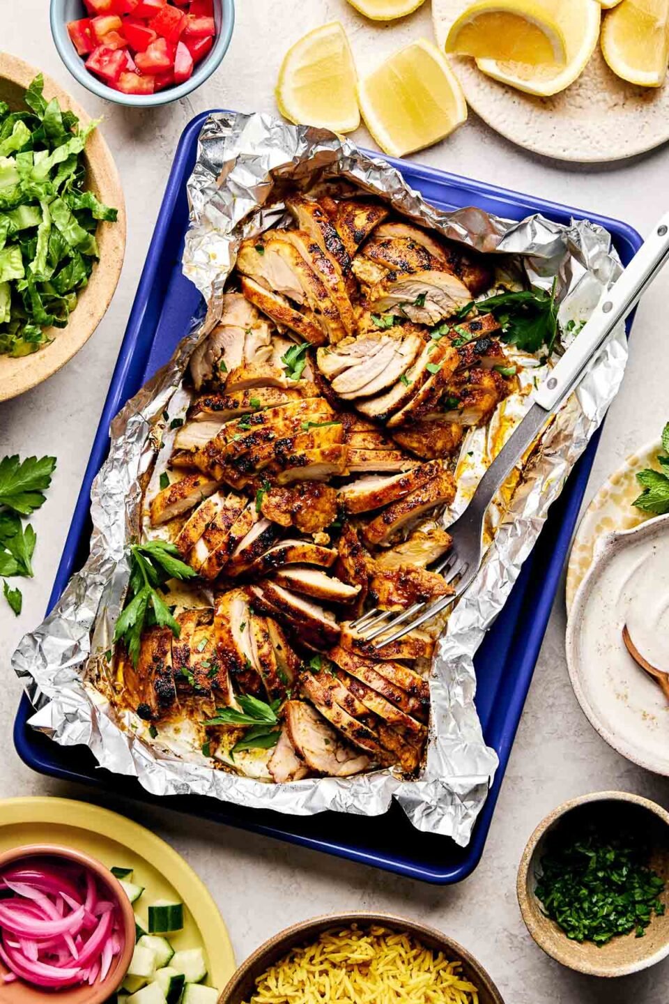 An overhead shot of sliced shawarma chicken thighs on a foil-lined blue sheet pan atop a white surface. The pan is surrounded by dishes of lemon wedges, diced tomatoes and cucumbers, shredded lettuce, pickled red onions, turmeric rice, shawarma sauce and fresh herbs.