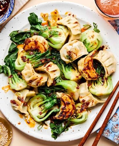 An overhead shot of cooked dumplings and bok choy drizzled with sauce on a white plate on a beige surface. A pair of chopsticks sits alongside the plate, along with dishes of sesame seeds and dumpling sauce.