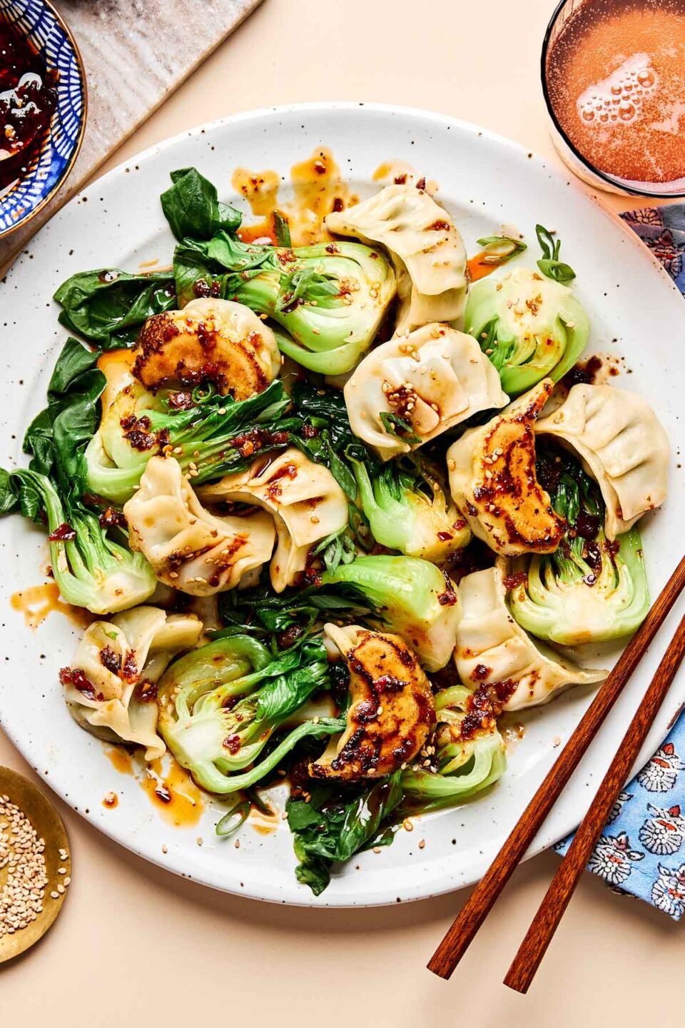 An overhead shot of cooked dumplings and bok choy drizzled with sauce on a white plate on a beige surface. A pair of chopsticks sits alongside the plate, along with dishes of sesame seeds and dumpling sauce.