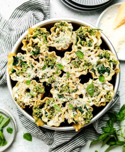 An overhead shot of a round pan of cooked lasagna rolls on a striped cloth atop a light grey surface. Grey plates, a plate of parmesan cheese and basil leaves sit beside it.