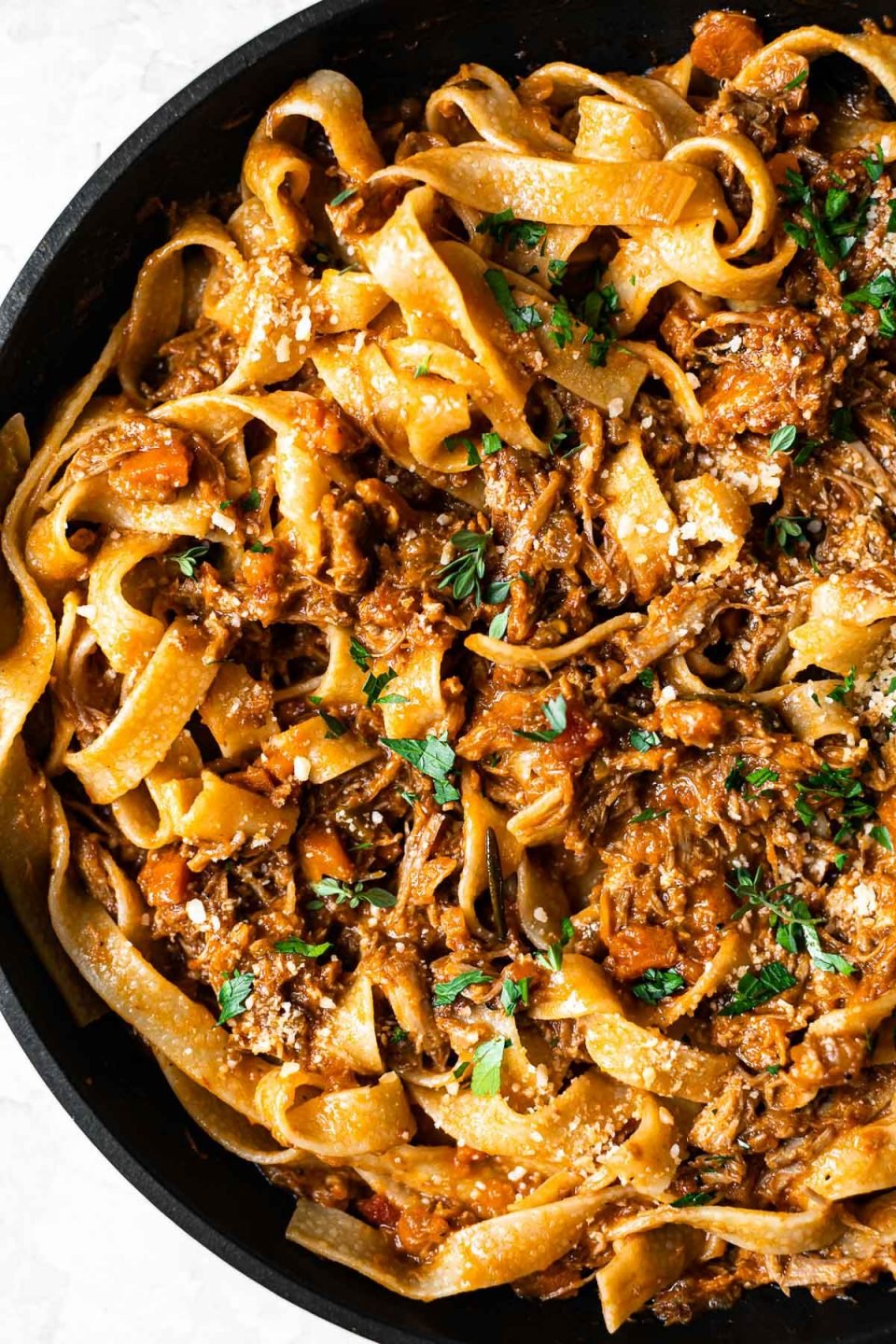 Close up of pork shoulder ragu tossed into pappardelle pasta in a skillet. The pasta is topped with fresh herbs.