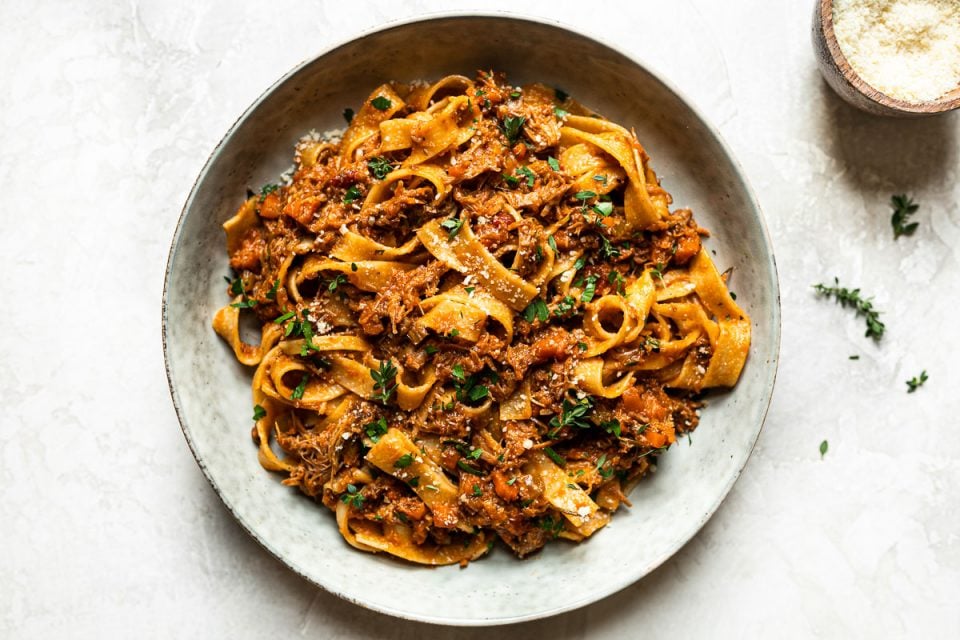 Pork ragu pappardelle shown in a large gray pasta bowl. The pasta is topped with fresh herbs. Surrounding the bowl is a small bowl of grated parmesan & some fresh thyme sprigs.