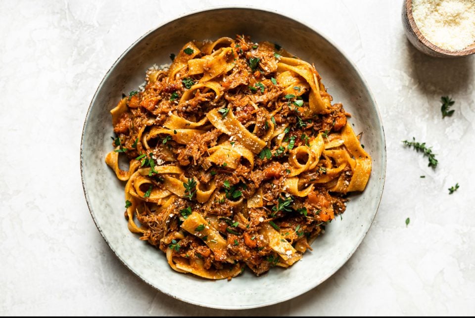 Pork ragu pappardelle shown in a large gray pasta bowl. The pasta is topped with fresh herbs. Surrounding the bowl is a small bowl of grated parmesan & some fresh thyme sprigs.