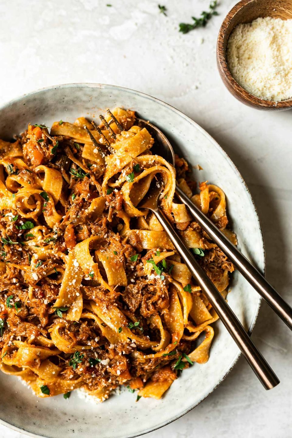 Pork ragu pappardelle shown in a large gray pasta bowl, with a fork & spoon nestled into the side of it. The pasta is topped with fresh herbs. Surrounding the bowl is a small bowl of grated parmesan & some fresh thyme sprigs.