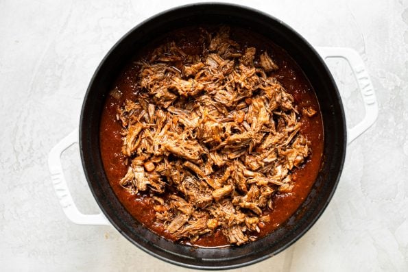 Shredded pork shoulder in tomato ragu sauce in a large white Dutch oven.