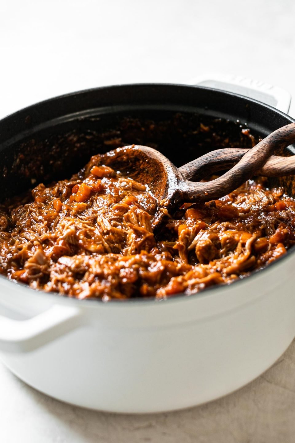 Pork ragu inside of in a large white Dutch oven. A wooden spoon is nestled into the pot for serving.