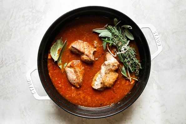 Browned pork shoulder with bay leaves & fresh herbs, nestled in tomato sauce in a large white Dutch oven.